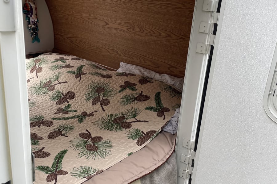 Inside an RV rental: a comfortable bed with a pinecone quilt, wood walls, and a small light. The door is open to the outside.