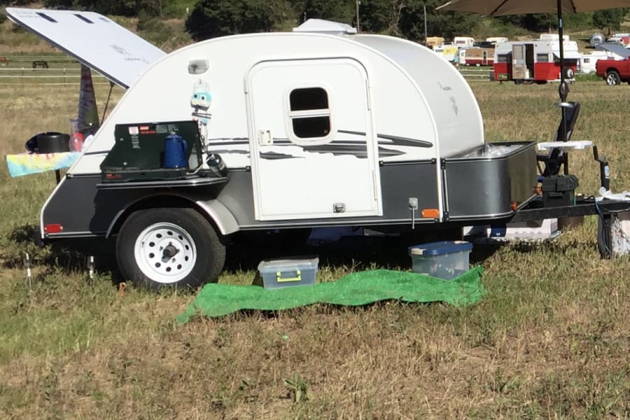 Teardrop RV trailer set up with an open kitchen hatch in a grassy field. Perfect for your next RV rental adventure!