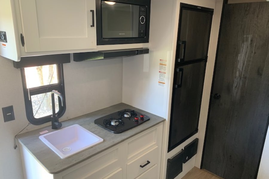 Modern RV rental kitchen with white cabinets, microwave, sink, 2-burner stove, and refrigerator.
