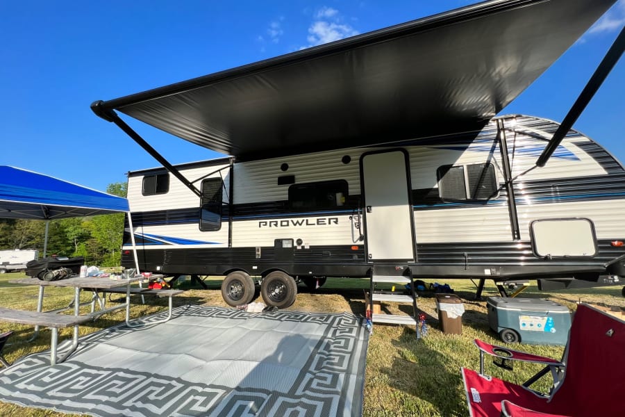 Sunny day RV rental setup. A travel trailer with an extended awning, outdoor mat, and picnic table at a grassy campsite.