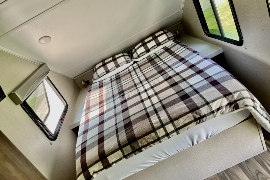 Serene RV rental bedroom featuring a comfy plaid bed, side windows showing greenery, and a ceiling light.