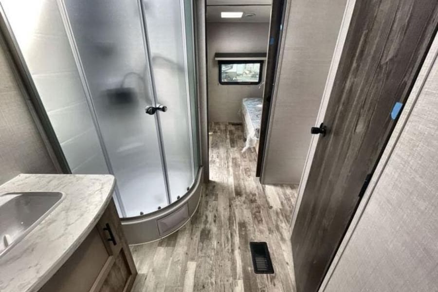 Inside an RV rental: a compact bathroom with a corner shower and sink, leading to the bedroom area on wood-look floors.