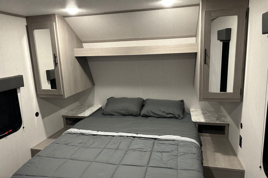 Modern RV rental bedroom: gray bed, compact nightstands with storage, and overhead lights for a cozy travel stay.