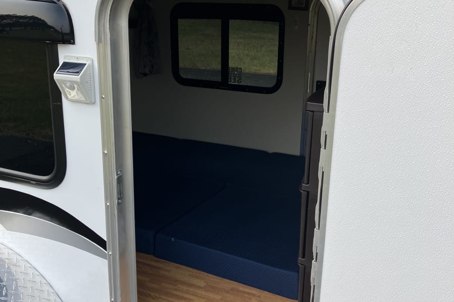 Compact white travel trailer RV rental interior with blue mattress, wood floor, solar light. Awning & solar panel visible.