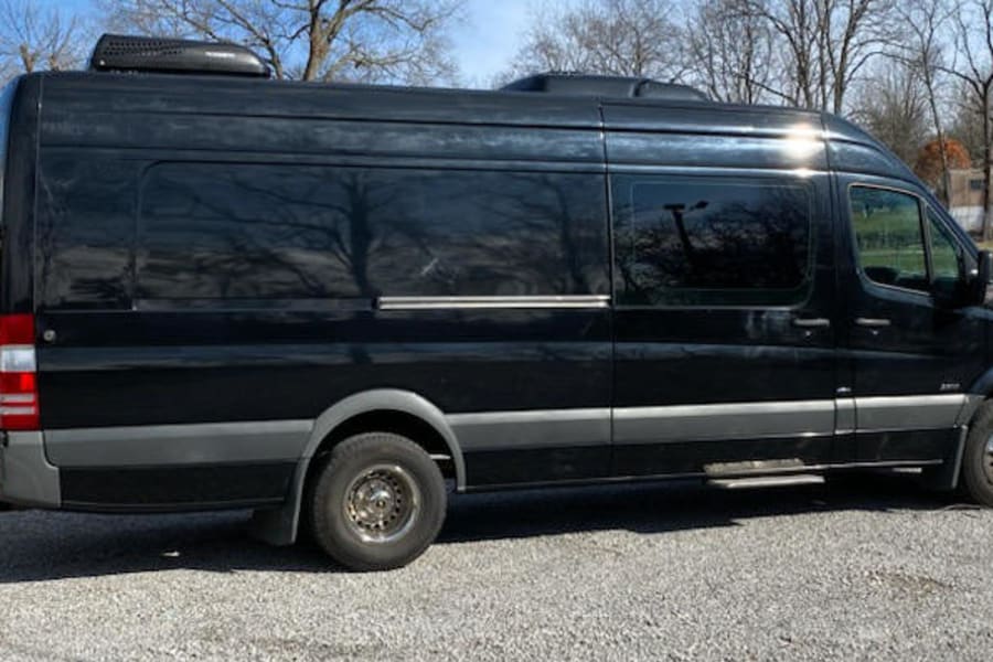 Black Mercedes-Benz Sprinter van, often used for RV rental, parked on a gravel lot next to other large vehicles.