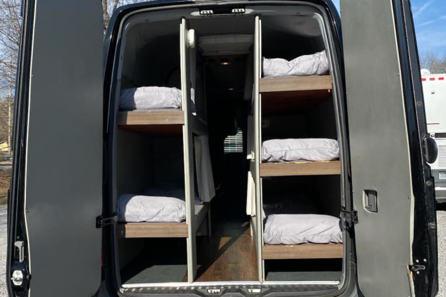 RV rental: Back of a black camper van with open doors reveals six bunk beds inside for mobile sleeping.