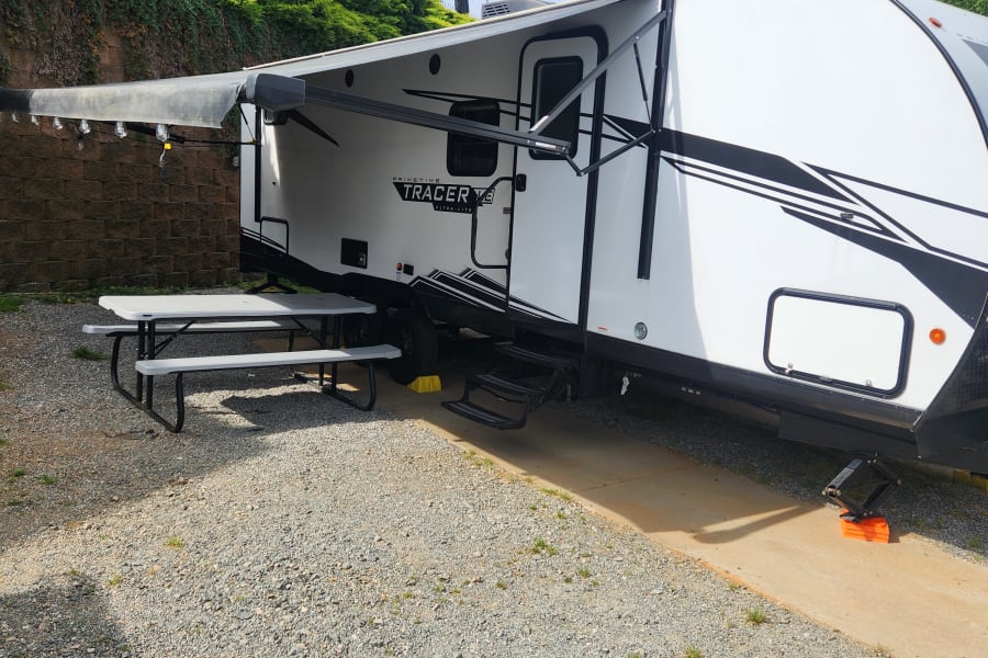 An RV rental, a white travel trailer, parked at a gravel campsite with a picnic table and extended awning. Perfect for camping.
