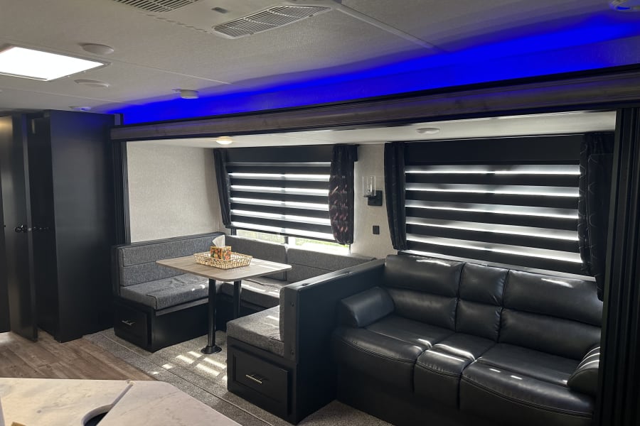 Spacious modern RV rental interior: black leather sofa, dinette with storage, and blue accent lighting.