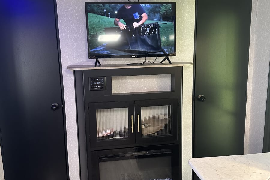Inside a modern RV rental, showing a well-equipped entertainment center with TV and electric fireplace, plus kitchen counter.