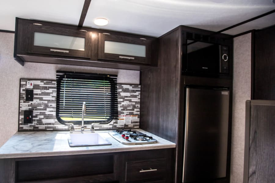 RV rental interior: modern compact kitchen with dark cabinets, mosaic backsplash, sink, stovetop, microwave, and fridge.