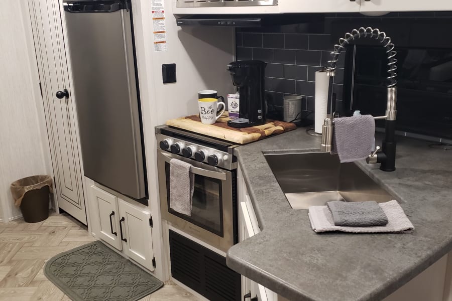 Modern RV rental kitchen with stainless steel microwave, sink, stove, and refrigerator. White cabinets and black hardware.