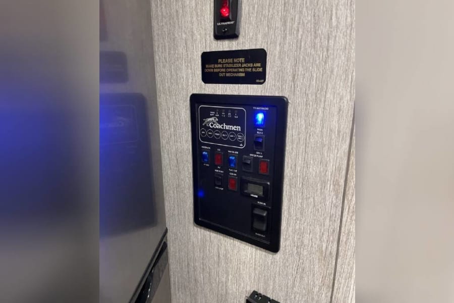 RV rental control panel on a light wall, displaying Coachmen features like water, lights, & slide-out controls with safety warnings.