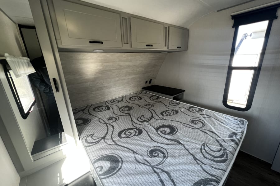 Inside a modern RV rental bedroom, featuring a patterned mattress, overhead cabinets, and natural light from windows.