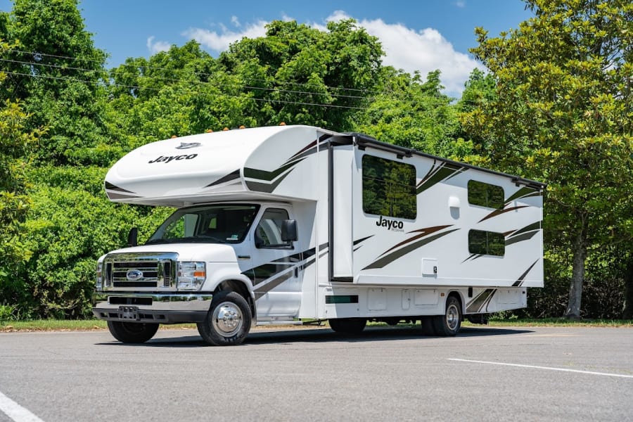 White Jayco Class C RV rental parked on asphalt, surrounded by green trees under a blue sky, ready for adventure.