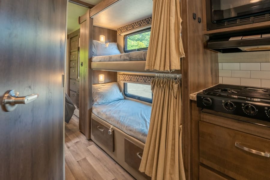 RV rental interior featuring cozy bunk beds with curtains, windows, and lamps, plus a compact kitchen with stovetop and microwave.