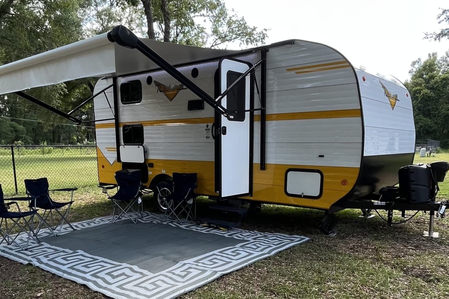 A vibrant yellow and white travel trailer RV rental with an awning extended over a mat and four folding chairs, perfect for camping.