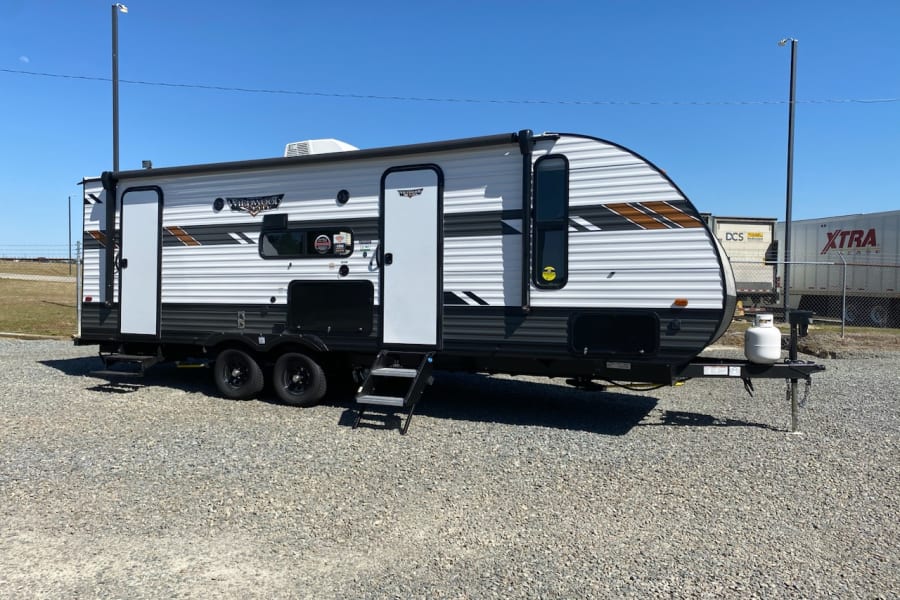 Spacious white & grey rv rental travel trailer parked on a gravel lot, with steps extended, under a clear blue sky.