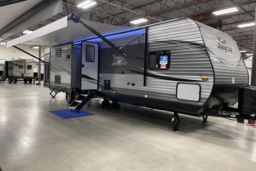 A grey Jayco travel trailer with an extended, blue-lit awning in a bright showroom. Ideal for your next RV rental adventure.