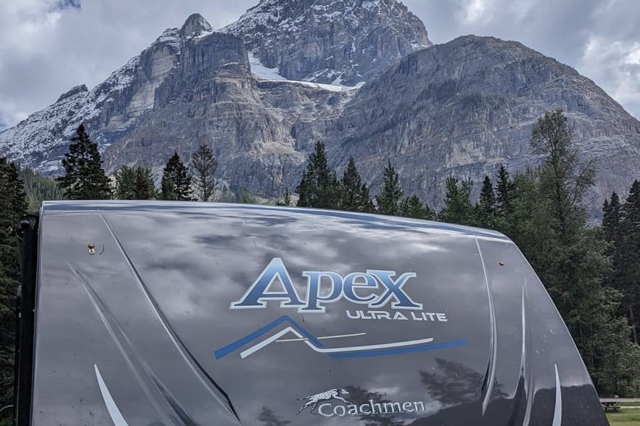 An Apex Ultra Lite rv rental before a magnificent, snow-capped mountain under cloudy skies. Perfect nature escape.