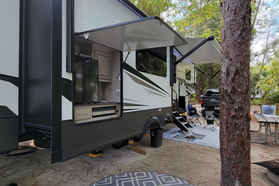Spacious RV rental set up at a shaded campsite, featuring an outdoor kitchen, awnings, and leisure area.
