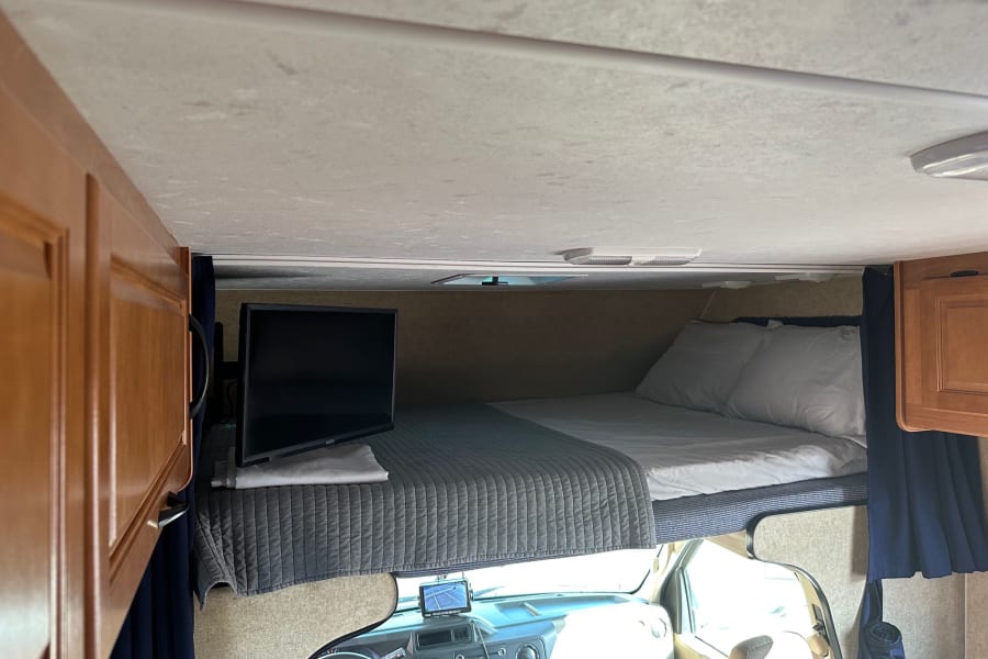 Inside an RV rental: a cozy cab-over bunk bed with a TV, pillows, and quilt, offering a comfy sleeping space above the driver's cabin.