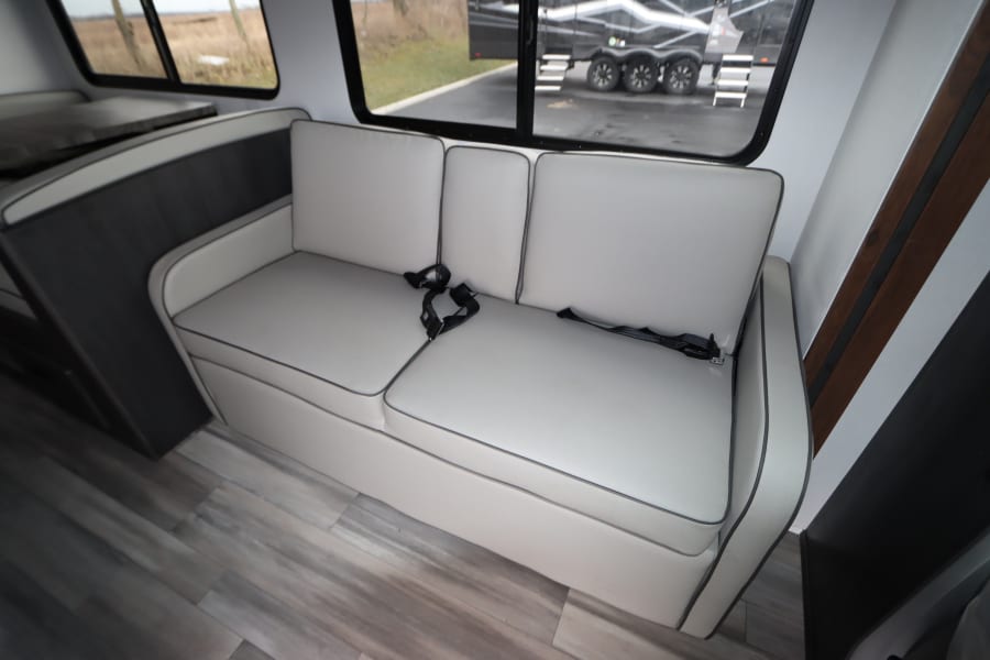 Spacious RV rental interior featuring a light gray sofa with seatbelts, wood flooring, and large windows.