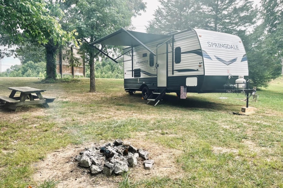 This RV rental is perfectly set up at a grassy campsite with a picnic table and fire pit, surrounded by trees.