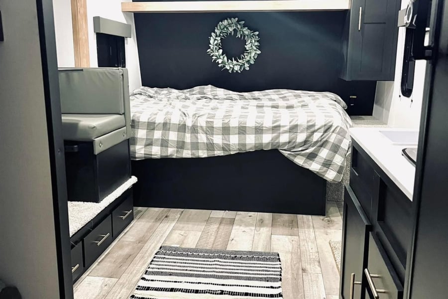 Modern RV rental interior with a bed, bench seating, and kitchenette. Features dark walls, plaid bedding, and light wood floors.