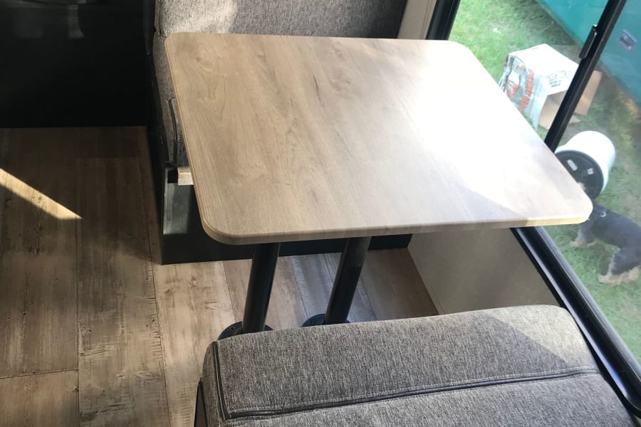 Experience a comfortable RV rental: dinette area with a light wood table and cozy grey seating, looking out.