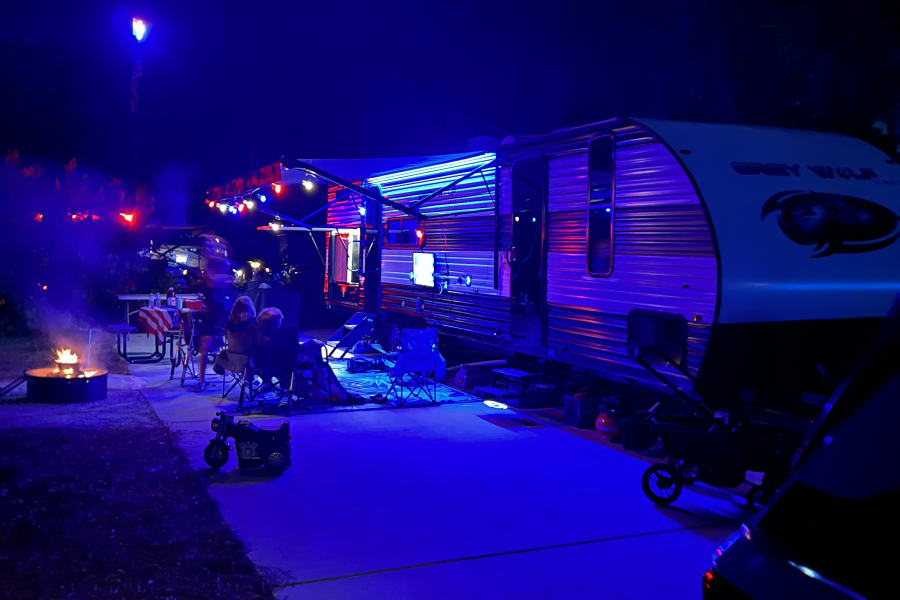 Relaxing night at an RV rental: Grey Wolf illuminated by vibrant blue and purple lights, people gathered by a campfire.