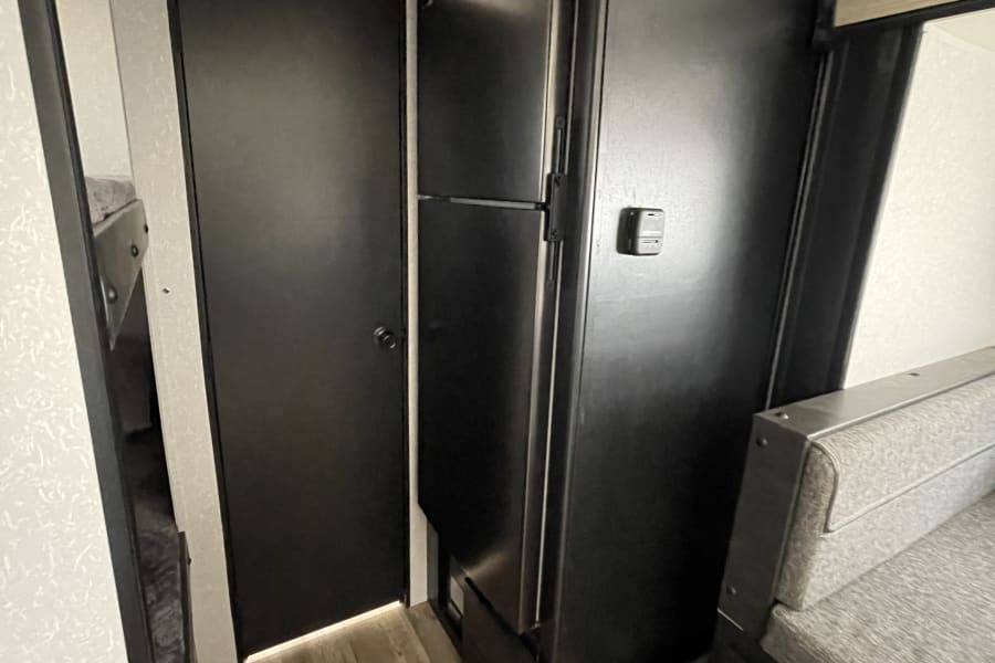 This RV rental interior features a black refrigerator, grey dinette seating with storage, and a door to bunk beds.
