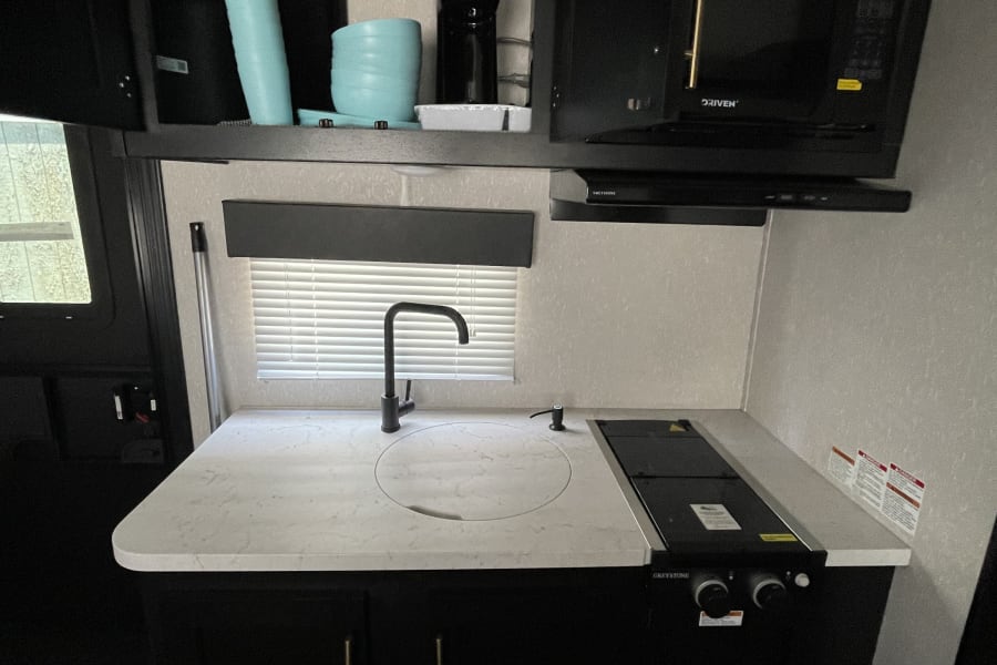 Compact and modern RV rental kitchen: Featuring a sink with a cover, cooktop, microwave, and overhead cabinets with blue dishes.