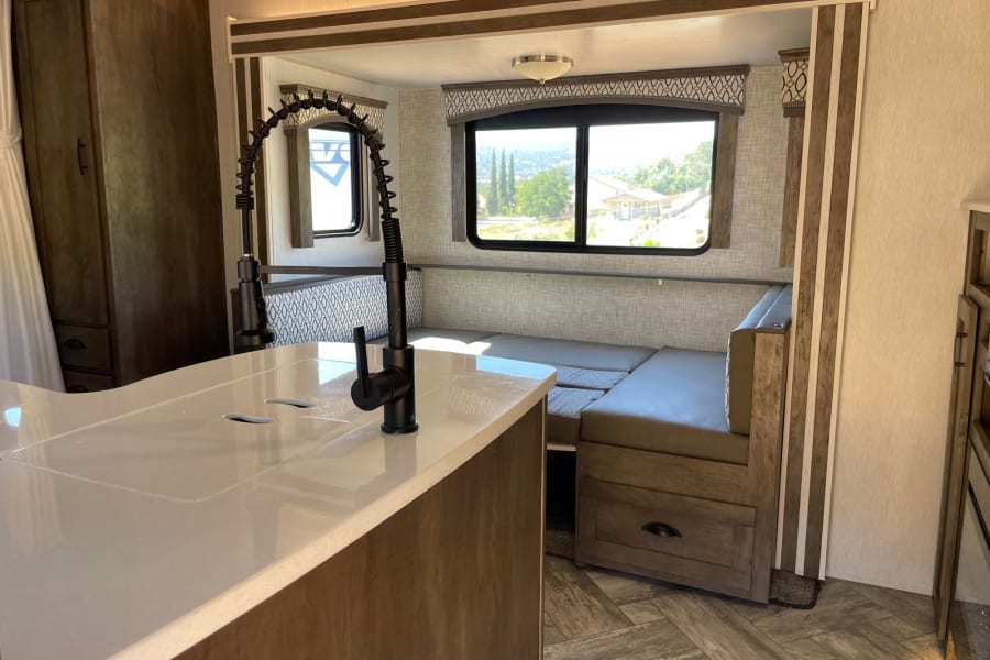 Bright RV rental interior: kitchen island, sleek faucet, dinette with scenic views from large windows. Modern travel trailer living.