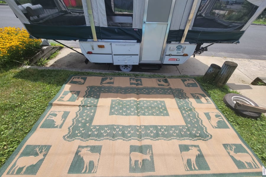 Pop-up RV rental set up on a concrete pad, with a large, patterned outdoor mat featuring deer designs on the grassy ground.
