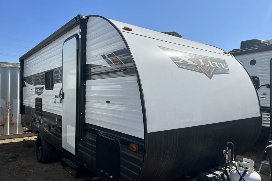 White and black travel trailer exterior, featuring an awning, door, and front propane tanks. Perfect for your RV rental.