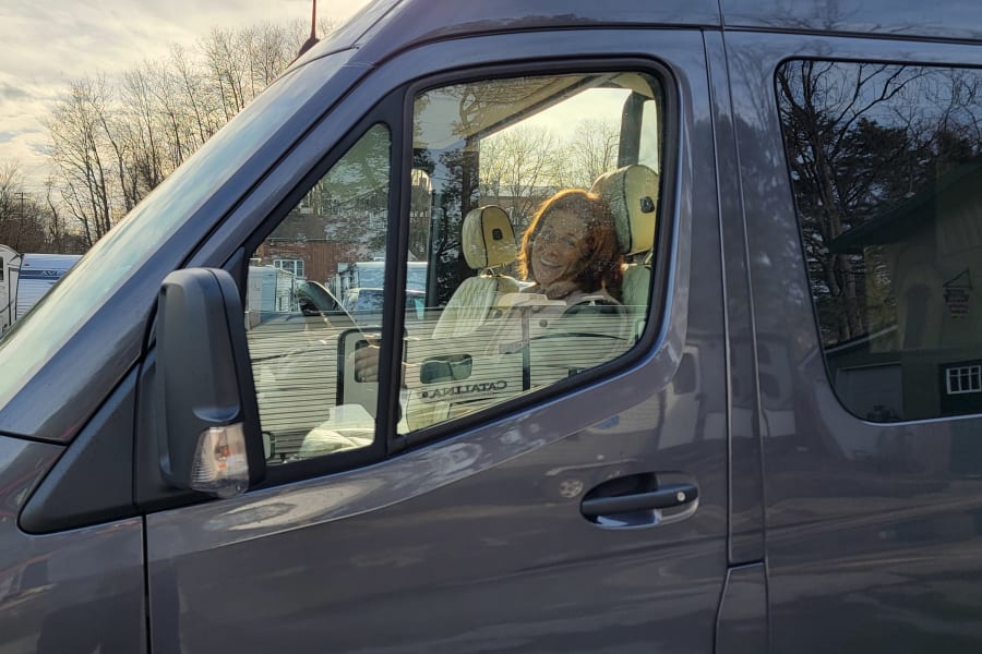 A grey RV rental camper van parked outdoors, showing a driver's side view with bright sun reflecting off the windows, a person visible inside.