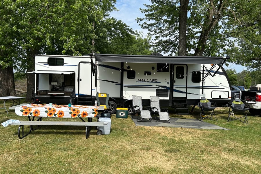 Enjoy outdoor living with this RV rental! A white travel trailer is set up at a sunny campsite with a picnic table and lounge chairs.