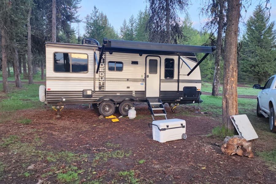 This RV rental offers a spacious camping experience amidst tall trees, with its awning extended for outdoor comfort.