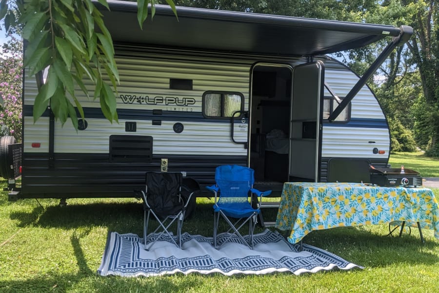 Peaceful RV rental campsite: travel trailer with awning, chairs, and griddle set up on a cozy outdoor rug.
