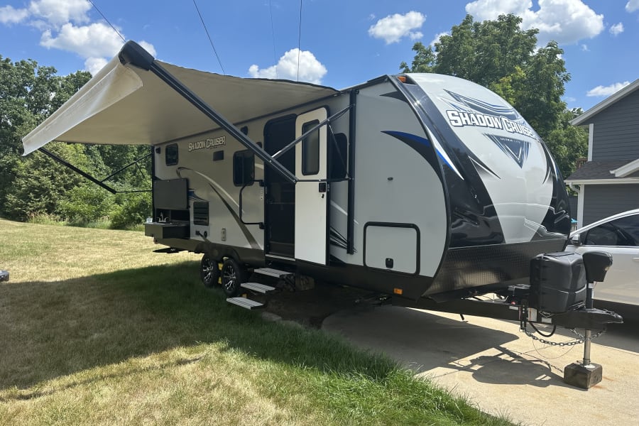 Sunny day highlights a Shadow Cruiser travel trailer, featuring an extended awning and outdoor kitchen, perfect for RV rental.