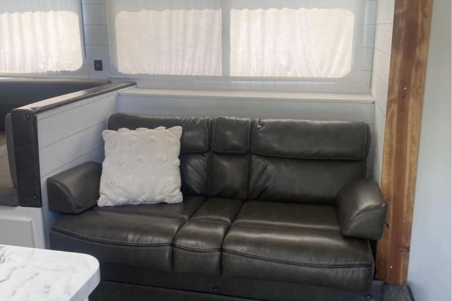Modern RV rental living space: cozy dark sofa, bright windows with shades, and rustic wood beam accents.