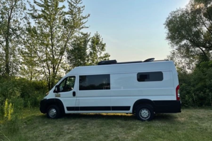 A sleek white camper van RV rental is parked on a grassy patch amidst green trees, inviting outdoor adventures.