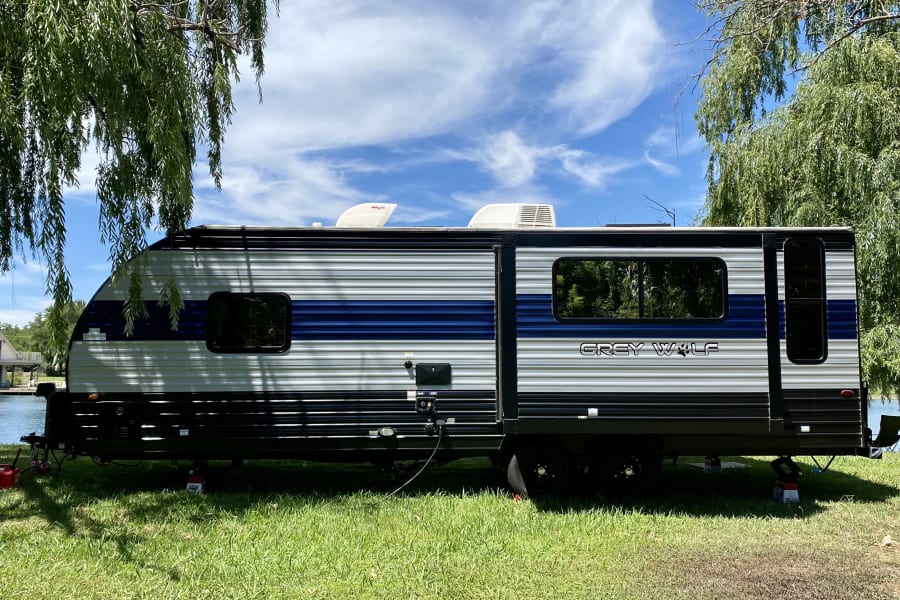 A Grey Wolf travel trailer, available as an rv rental, is parked lakeside, shaded by a willow, ready for adventure.