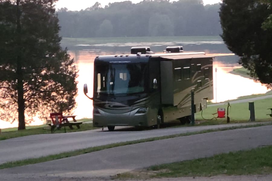 An RV rental is parked at a scenic lakefront campsite, slide-outs extended. Golden light reflects beautifully on the water.