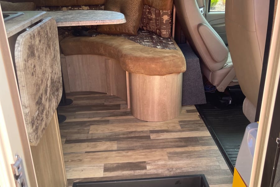 This rv rental interior features a cozy dining area with a U-shaped couch, table, wood-look floor, and a view into the cockpit.