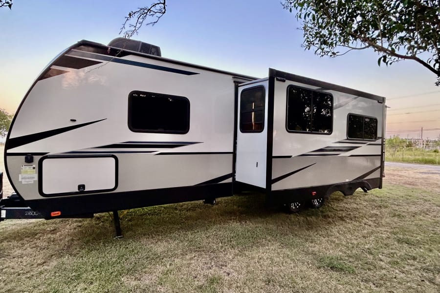 This spacious RV rental, a light-colored travel trailer with its side expanded, is parked on green grass under a clear sky.