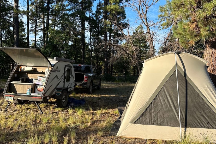 Scenic campsite with an RV rental (teardrop camper), tent, and fire pit in a pine forest.