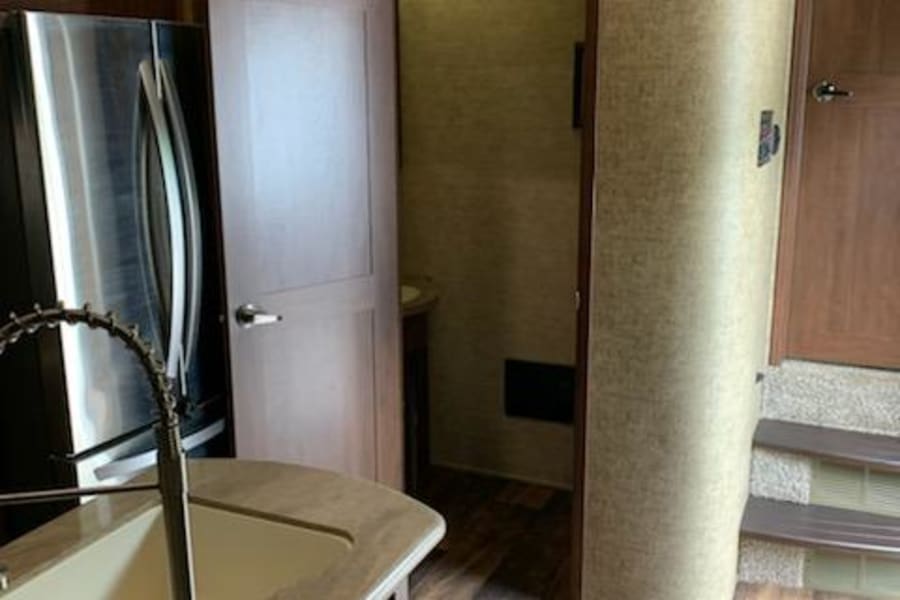 RV rental interior: Kitchen island with sink, stainless fridge, and open door to the private bath. Wood-look floors.