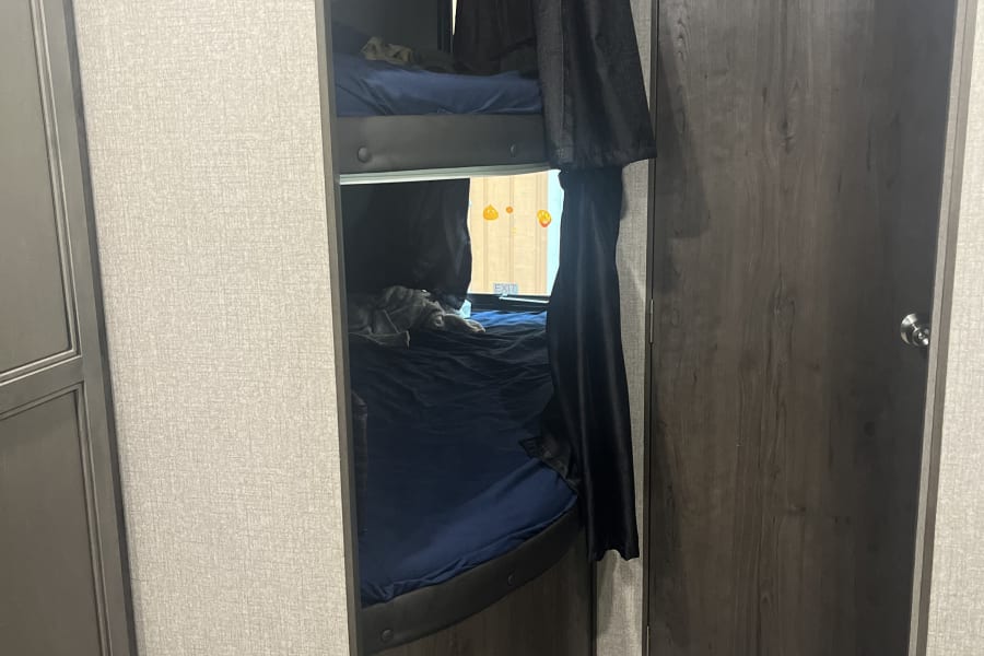 An RV rental features two bunk beds with dark privacy curtains, a window, floor vent, and a door with coat hooks.