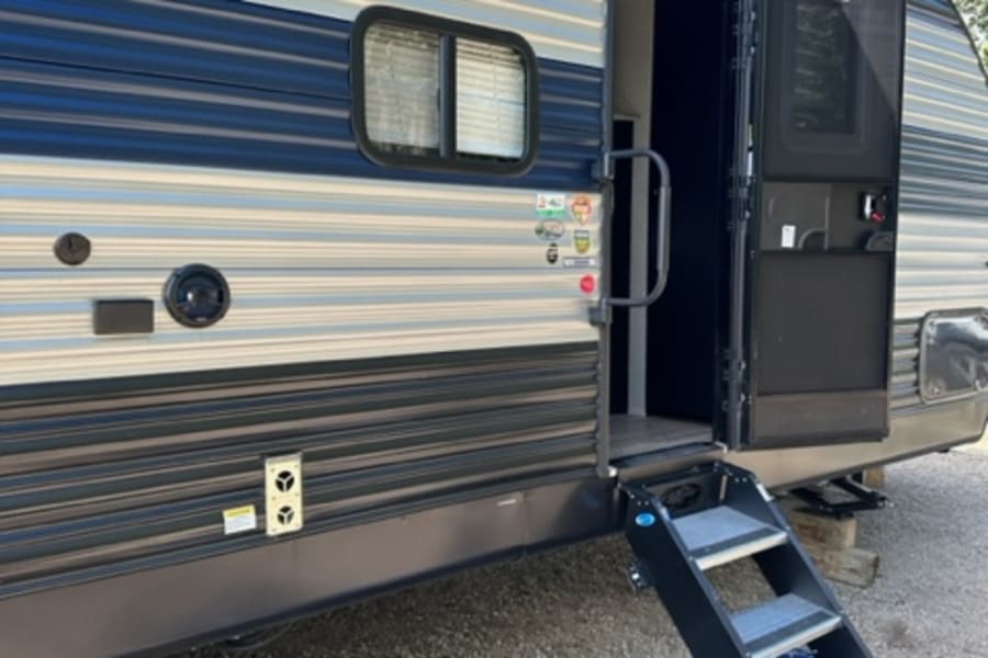 RV rental showing a striped travel trailer with its door open, revealing interior steps leading to an outdoor rug.